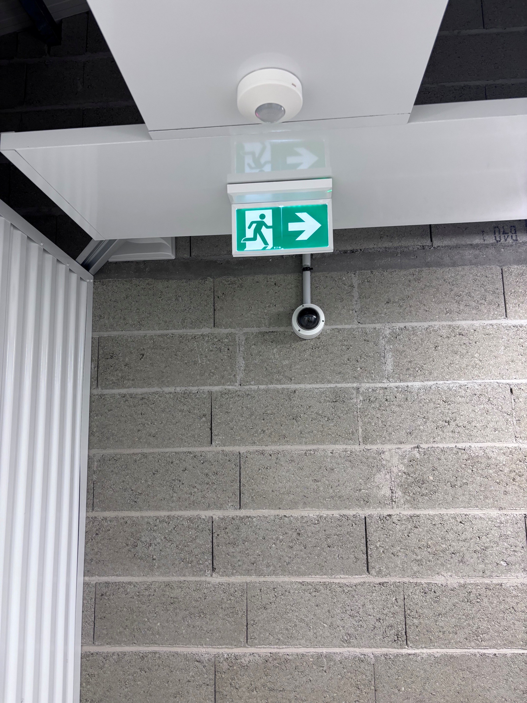 Dispositifs de sécurité dans un bâtiment de stockage : un panneau de sortie de secours lumineux vert et une caméra de vidéosurveillance.